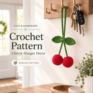 May include: A handmade crochet cherry hanger decor featuring two bright red spherical cherries attached to green stems and leaves. The text reads CUTE & HANDMADE, Crochet Pattern, Cherry Hanger Decor, and ENGLISH PATTERN. It hangs from a wooden hook.