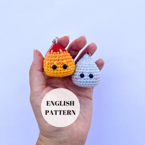 May include: Two crocheted keychains, one orange and one light blue, each with a smiling face. The orange keychain has red accents and the light blue one has pink accents. The keychains are held in a hand. The image includes the text "ENGLISH PATTERN".