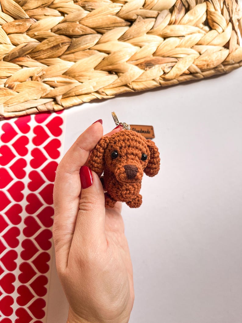 May include: A brown crocheted dog keychain with black eyes and a black nose. The keychain is held in a hand with red nail polish. The tag on the keychain says "Handmade".