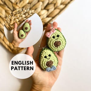 Puede incluir: Dos llaveros de aguacate tejidos a crochet, uno con un lazo rosa y el otro con un lazo azul. Los aguacates tienen caras sonrientes y semillas marrones. El texto "ENGLISH PATTERN" es visible en la imagen.