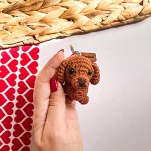May include: A brown crocheted dachshund dog keychain with black eyes and a black nose. The keychain is attached to a silver key ring and has a small brown leather tag that says "Handmade".
