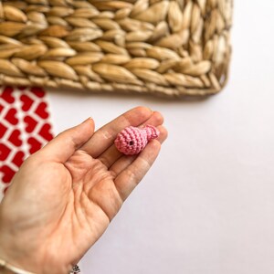 Puede incluir: Un peque&ntilde;o adorno de coraz&oacute;n de color rosa tejido a crochet. El adorno se sostiene en la mano de una persona.