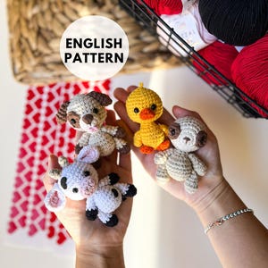 Könnte beinhalten: Eine Hand hält vier gehäkelte Amigurumi-Spielzeuge: einen braunen und weißen Hund, ein gelbes Küken, einen braunen Bären und eine schwarz-weiße Kuh. Die Spielzeuge sind klein und süß, perfekt für ein Kinderzimmer.