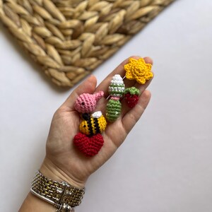 Puede incluir: Una mano sosteniendo una colecci&oacute;n de peque&ntilde;os art&iacute;culos tejidos a crochet, incluyendo una abeille, un coraz&oacute;n, una fresa, un sol y una flor. Los art&iacute;culos son todos de colores brillantes y hechos con hilo.