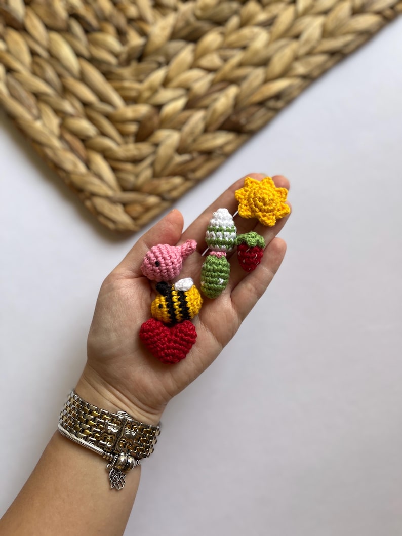 Puede incluir: Una mano sosteniendo una colecci&oacute;n de peque&ntilde;os art&iacute;culos tejidos a crochet, incluyendo una abeille, un coraz&oacute;n, una fresa, un sol y una flor. Los art&iacute;culos son todos de colores brillantes y hechos con hilo.