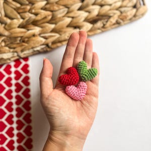 May include: Three small crocheted hearts in different colors: pink, red, and green. The hearts are held in a person's hand.