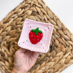 May include: A pink crocheted square with a red and green strawberry design in the centre.