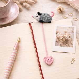 May include: A handmade crocheted opossum bookmark with a pink heart. The grey and white opossum sits on an open notebook, next to a pink and white striped pen. A cup of coffee and a photo are also visible.