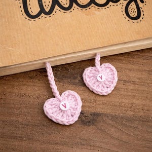 May include: Two pink crocheted heart bookmarks with white buttons and a braided cord, resting on a wooden surface. The word "tweety!" is written on a brown book.