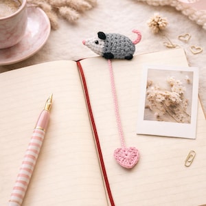 May include: A handmade crocheted opossum bookmark with a pink heart, placed on an open notebook. The opossum is grey with black details and a pink tail. A pink and white striped pen and a coffee cup are also in view.