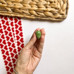 Puede incluir: Un peque&ntilde;o cactus verde tejido a crochet con flores rosas en la parte superior. El cactus est&aacute; siendo sostenido en una mano.