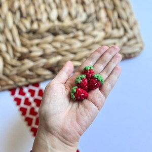 Puede incluir: Una mano sosteniendo cuatro fresas peque&ntilde;as de ganchillo. Las fresas son rojas con hojas verdes.
