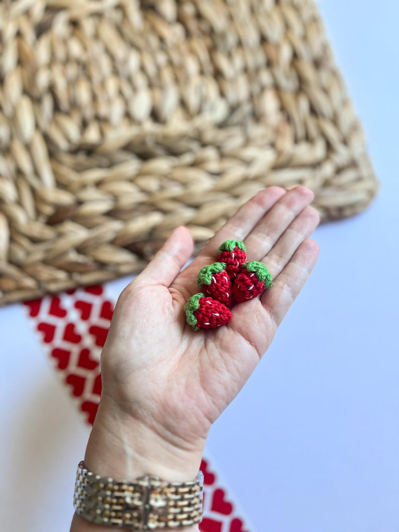 Puede incluir: Una mano sosteniendo cuatro fresas peque&ntilde;as de ganchillo. Las fresas son rojas con hojas verdes.
