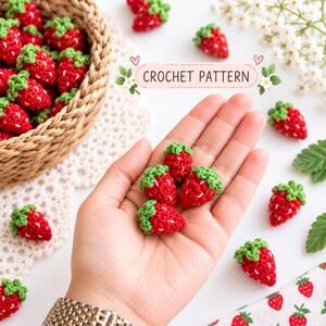 May include: Small red crocheted strawberries with green tops and white embroidered seeds. A woven basket contains several strawberries, while others are scattered around. The text CROCHET PATTERN is displayed in a decorative frame.