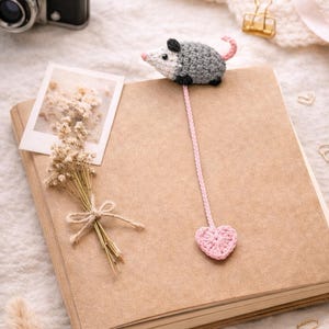 May include: A crochet bookmark with a grey and white mouse and a pink heart. The bookmark is on a brown notebook. A vintage camera and dried flowers are also visible. The bookmark is a charming, handmade item.