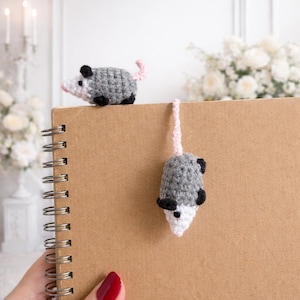 May include: Crocheted opossum bookmark and decorative figurine. The opossums are grey with black accents and pink tails. One sits atop a spiral-bound notebook, while the other hangs from the top. The notebook is brown.