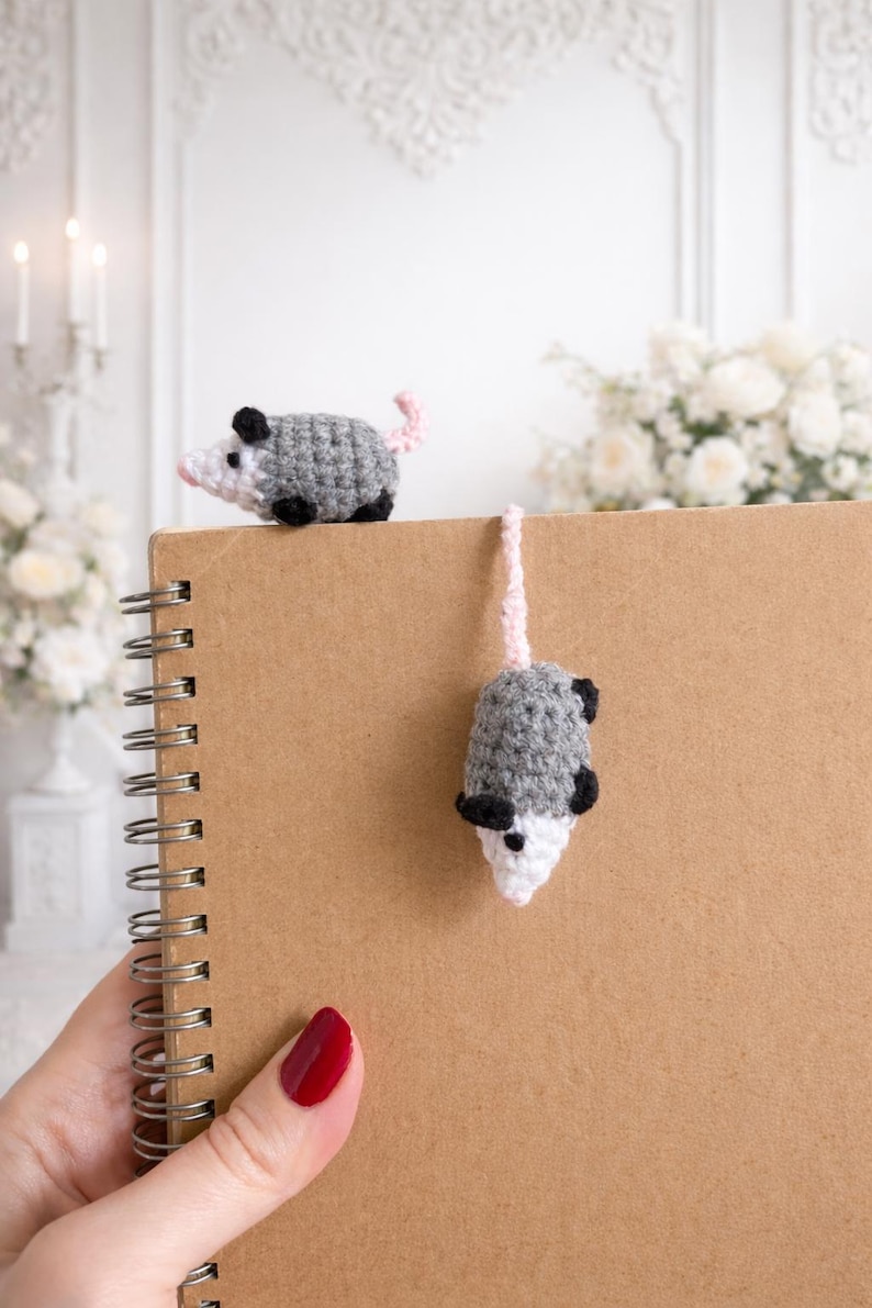 May include: Crocheted opossum bookmark and decorative figurine. The opossums are grey with black accents and pink tails. One sits atop a spiral-bound notebook, while the other hangs from the top. The notebook is brown.
