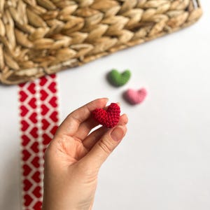 May include: A hand holding a small red crocheted heart. There are two other crocheted hearts in the background, one green and one pink.