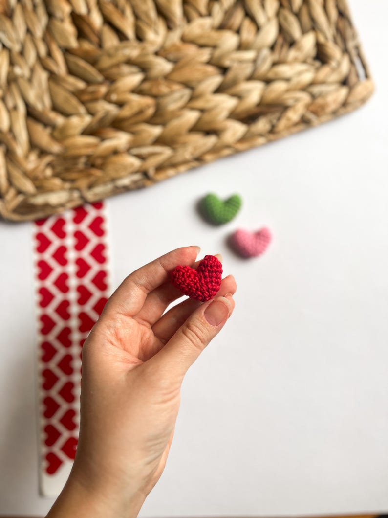 May include: A hand holding a small red crocheted heart. There are two other crocheted hearts in the background, one green and one pink.