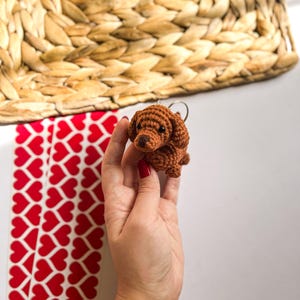 May include: A brown crocheted dachshund keychain with a key ring. The keychain is being held by a hand with red nail polish.