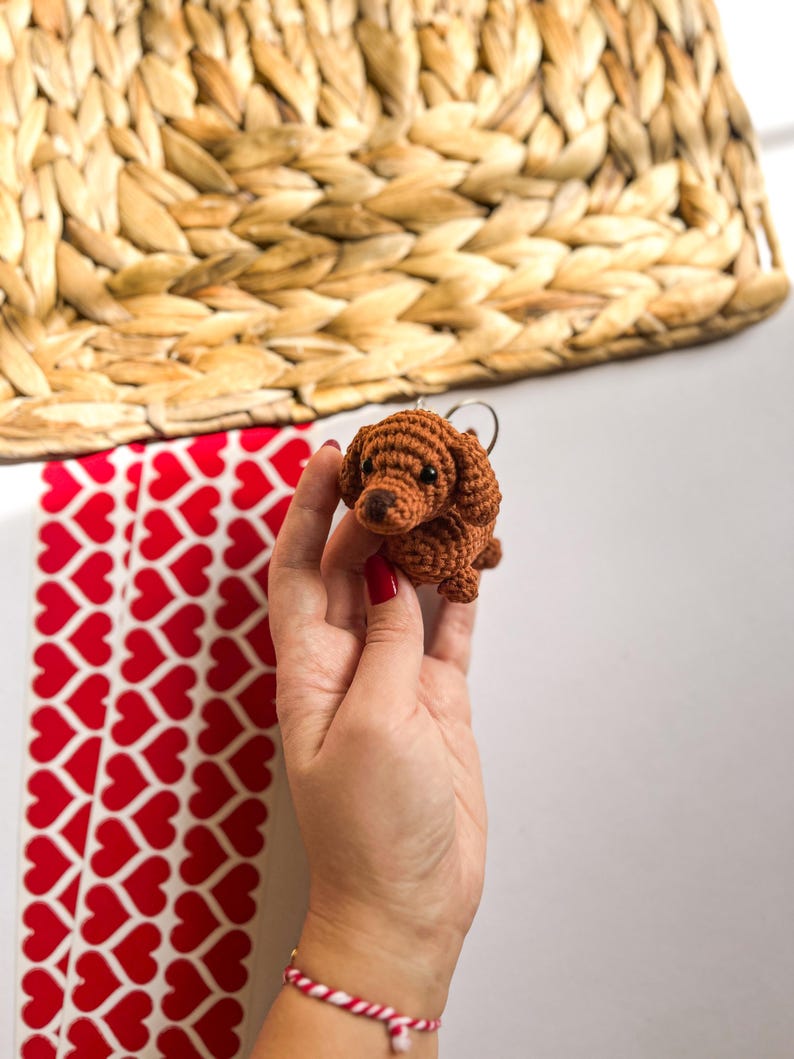 May include: A brown crocheted dachshund keychain with a key ring. The keychain is being held by a hand with red nail polish.