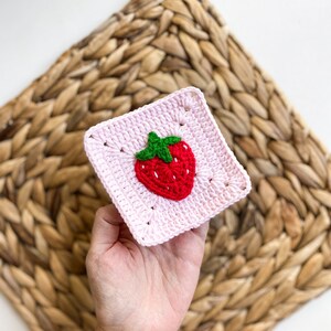 May include: A pink crocheted square with a red and green strawberry design in the centre.