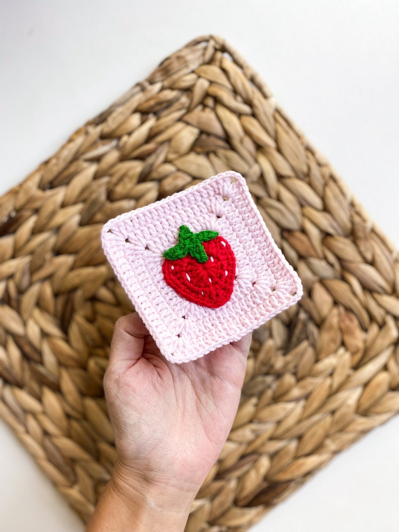 May include: A pink crocheted square with a red and green strawberry design in the centre.