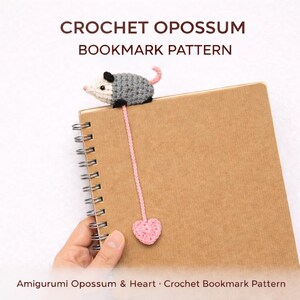 May include: A crochet opossum bookmark pattern. The bookmark features a grey and white amigurumi opossum with a pink tail, attached to a pink cord ending in a crocheted heart. The text "CROCHET OPOSSUM BOOKMARK PATTERN" is at the top.