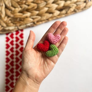 Puede incluir: Tres corazones de ganchillo en rosa, rojo y verde. Los corazones se sostienen en una mano.