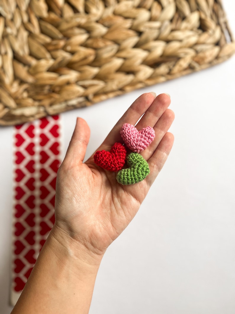 Puede incluir: Tres corazones de ganchillo en rosa, rojo y verde. Los corazones se sostienen en una mano.