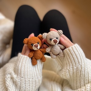 May include: Two small crocheted teddy bears, one brown and one beige, are held in the hands of a person wearing a white knitted sweater.
