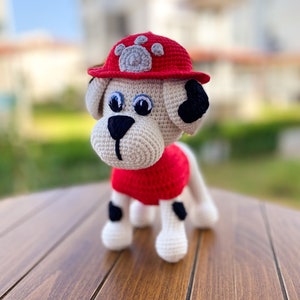 May include: A crocheted stuffed dog wearing a red firefighter hat with a paw print on the front. The dog has black spots and white fur.
