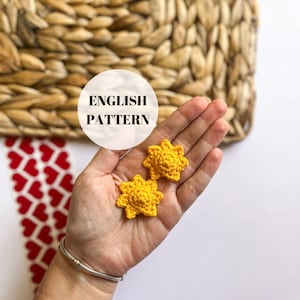 May include: Two crocheted yellow sun shapes, with a raised center, are held in a hand. The text "ENGLISH PATTERN" is on a white circle behind the hand.