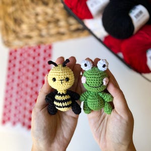 Puede incluir: Dos animales de peluche tejidos a crochet, una abeja y una rana, se sostienen en las palmas de dos manos. La abeja es amarilla y negra con ojos negros y una peque&ntilde;a sonrisa. La rana es verde con ojos blancos y un ligero rubor rosado en las mejillas.
