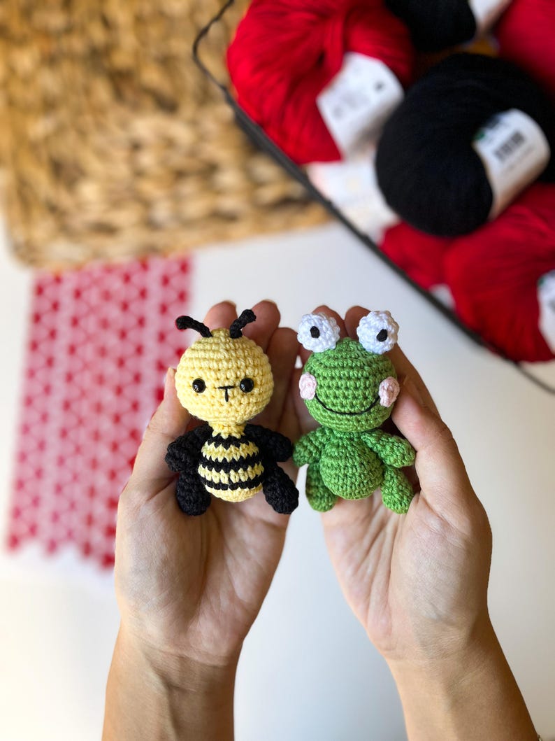 Puede incluir: Dos animales de peluche tejidos a crochet, una abeja y una rana, se sostienen en las palmas de dos manos. La abeja es amarilla y negra con ojos negros y una peque&ntilde;a sonrisa. La rana es verde con ojos blancos y un ligero rubor rosado en las mejillas.
