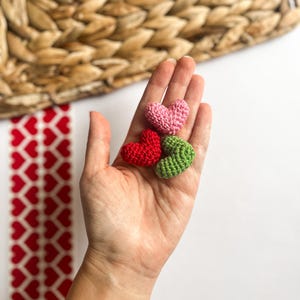 May include: Three crocheted hearts in different colors: pink, red, and green. The hearts are held in a hand.