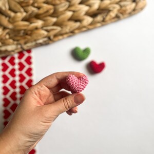 May include: A pink crocheted heart held in a hand. There are two other crocheted hearts in the background, one green and one red.