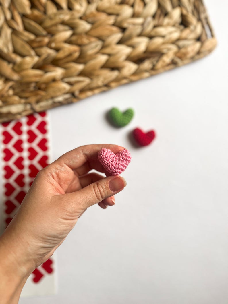 May include: A pink crocheted heart held in a hand. There are two other crocheted hearts in the background, one green and one red.