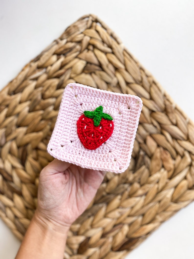 May include: A pink crocheted square with a red and green strawberry design in the centre.