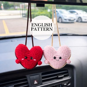 Op de afbeelding: Twee gehaakte hartvormige auto-hangers. Eén is rood, de andere is roze. Beide hebben zwarte ogen, kleine glimlachjes en kleine armpjes. De hangers hangen aan bruine en beige koorden. De tekst "ENGLISH PATTERN" staat in een witte cirkel.