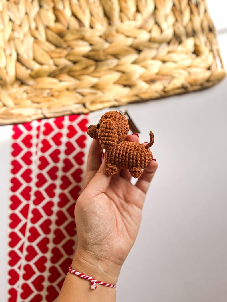 May include: A small brown crocheted dachshund toy held in a hand. The toy has a long body and short legs.