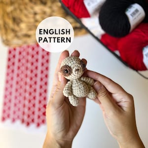 May include: A crocheted sloth amigurumi toy with brown eyes and a beige body. The toy is being held by a person's hands.