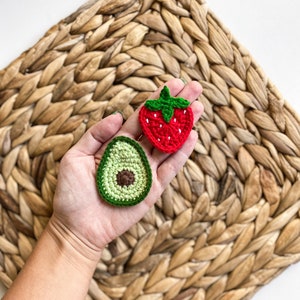 May include: A hand holding two crocheted food items: a green avocado and a red strawberry. The items are on a woven straw background.