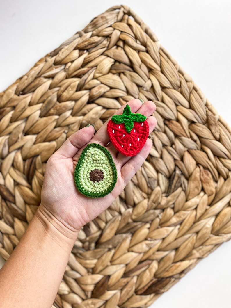 May include: A hand holding two crocheted food items: a green avocado and a red strawberry. The items are on a woven straw background.