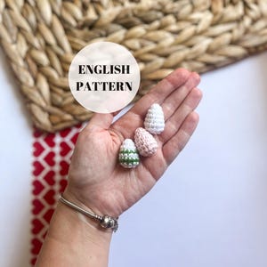May include: Three crocheted Easter eggs in different colors: white, pink, and green with white stripes. The eggs are held in a hand, with the text "ENGLISH PATTERN" in a white circle on the background.