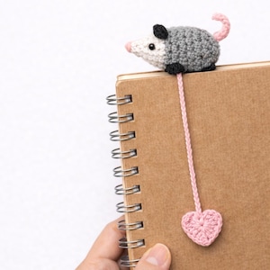 May include: A handmade crochet bookmark featuring a grey and white opossum with a pink nose and tail. The bookmark has a pink cord that ends in a pink heart. The opossum is perched on a brown spiral notebook.