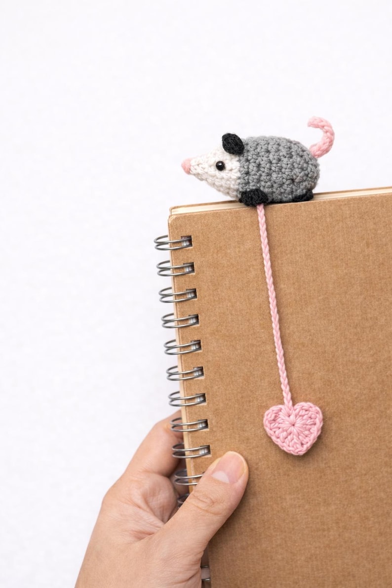May include: A handmade crochet bookmark featuring a grey and white opossum with a pink nose and tail. The bookmark has a pink cord that ends in a pink heart. The opossum is perched on a brown spiral notebook.