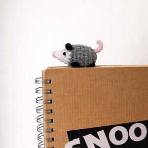 May include: A crocheted opossum in grey, white, black, and pink sits on a spiral-bound notebook. The notebook is brown with the word "SNOO" in white letters on a black rectangle.