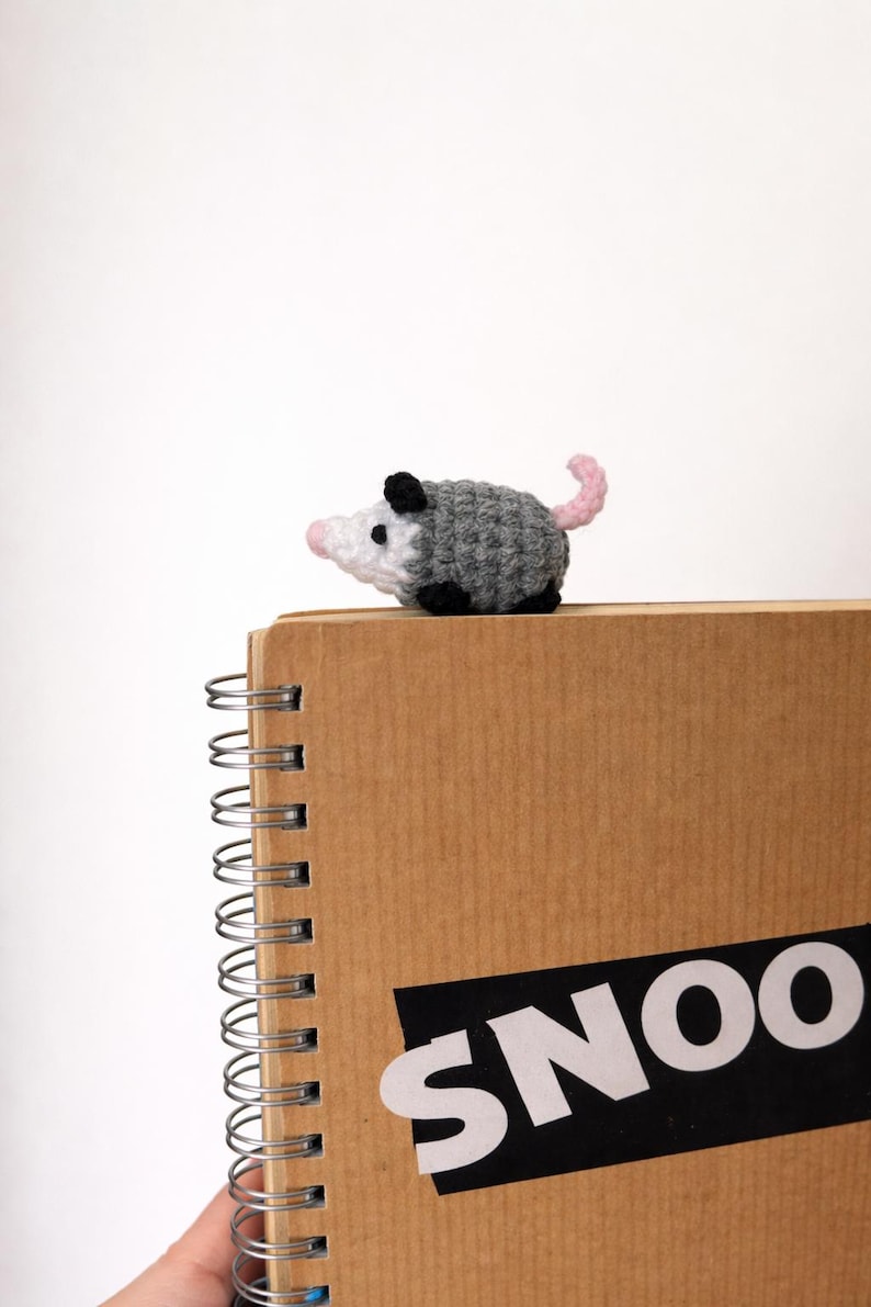 May include: A crocheted opossum in grey, white, black, and pink sits on a spiral-bound notebook. The notebook is brown with the word "SNOO" in white letters on a black rectangle.