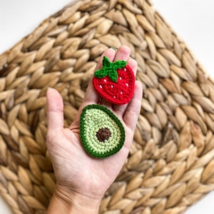May include: A hand holding two crocheted food items: a red strawberry and a green avocado. The items are on a woven straw background.